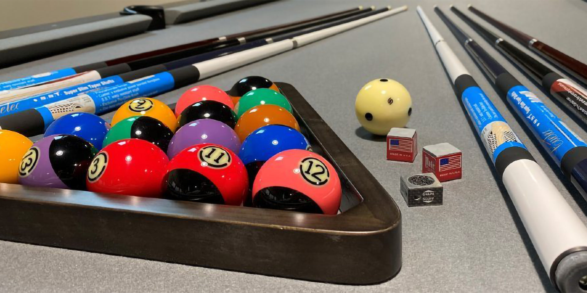 A set of pool balls in a triangle rack next to several pool cues and chalkers